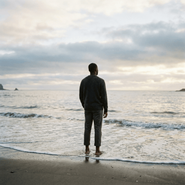 Man standing barefoot at the ocean shoreline beneath cloudy skies